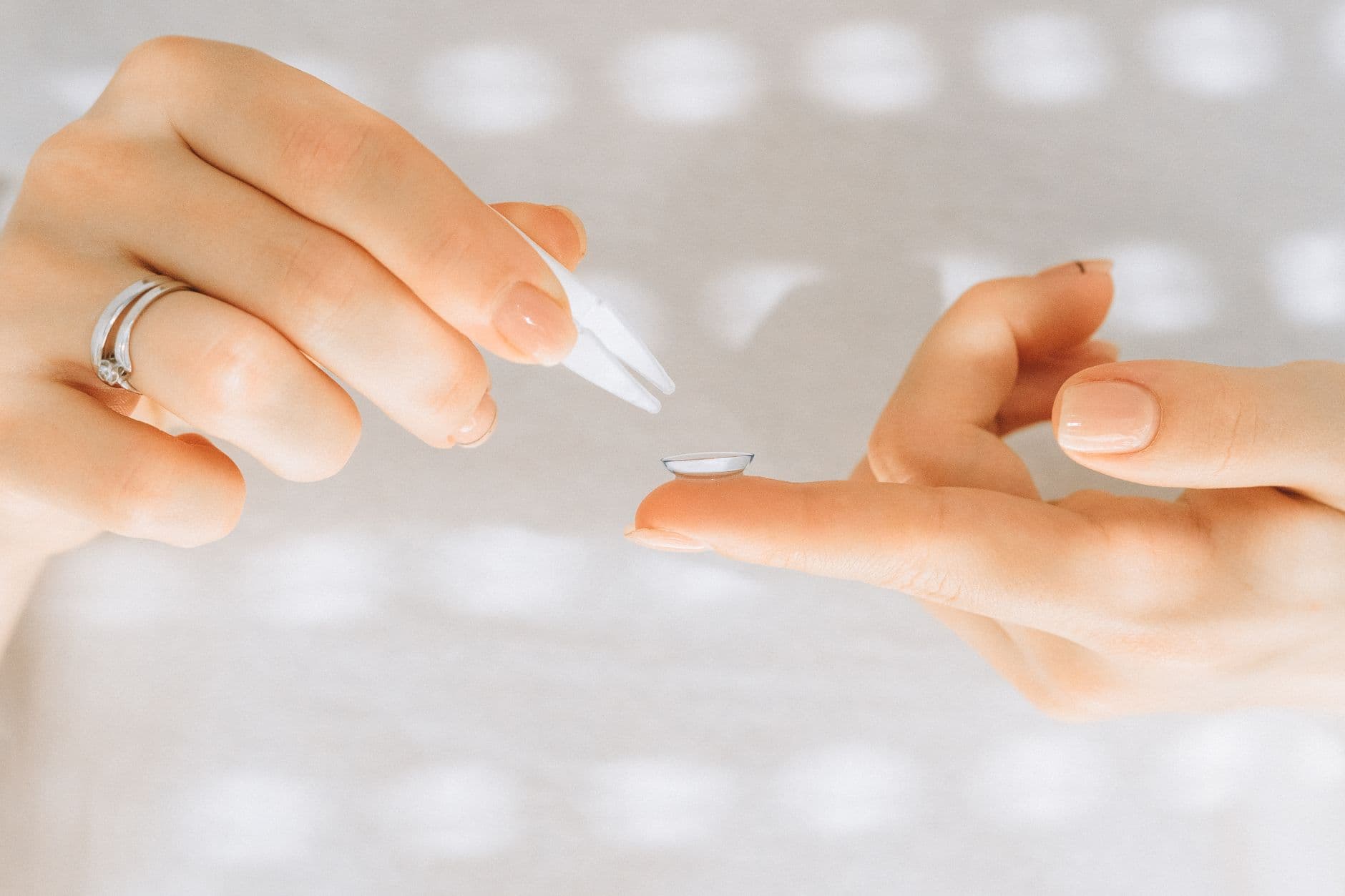 Manos sujetando una lentilla transparente en la yema del dedo junto a unas pinzas para lentes de contacto