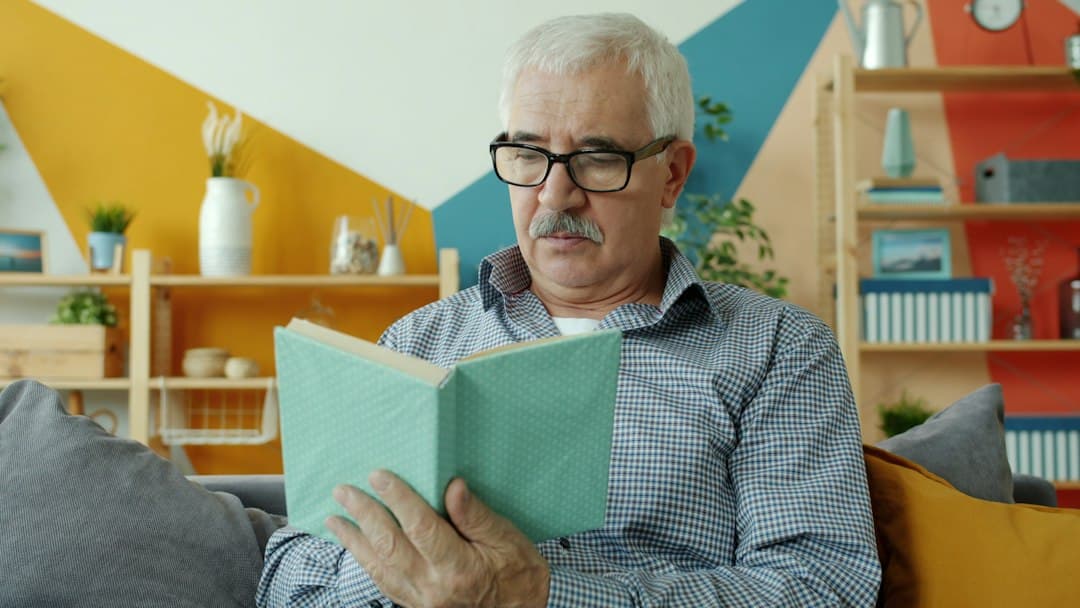 Hombre mayor con gafas graduadas leyendo un libro en el sofá de su casa