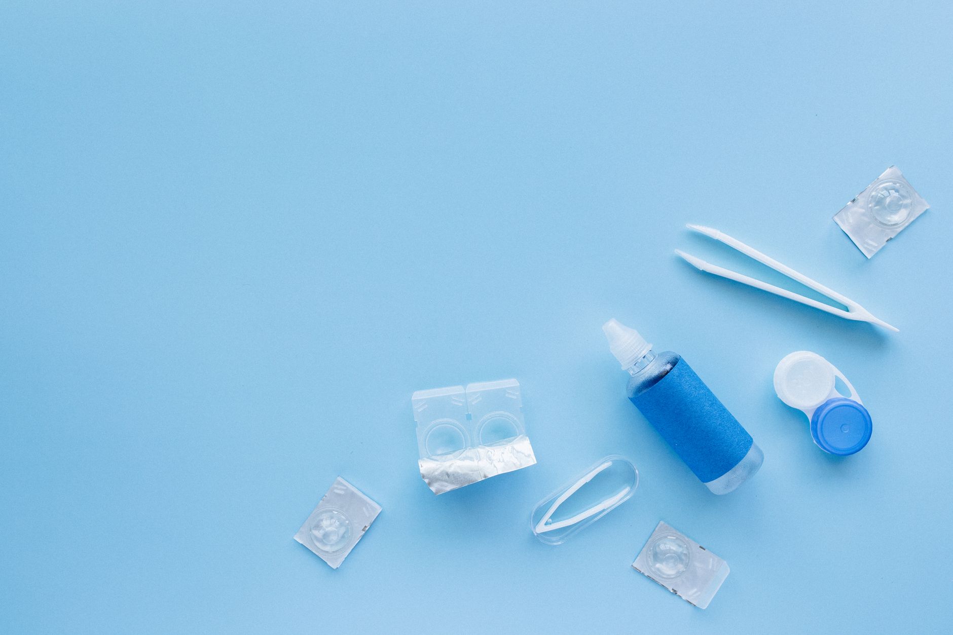 Blísteres de lentillas desechables junto a un estuche, unas pinzas y un bote de solución sobre fondo azul