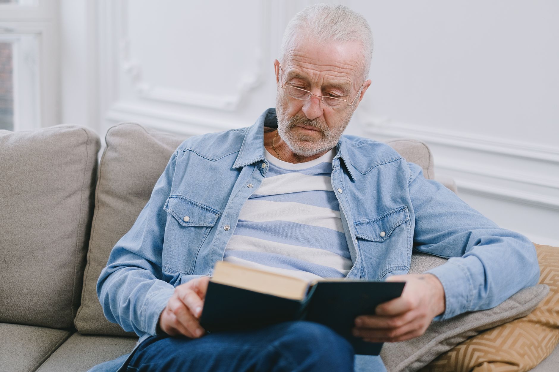 Hombre mayor con gafas de cerca leyendo un libro sentado en el sofá